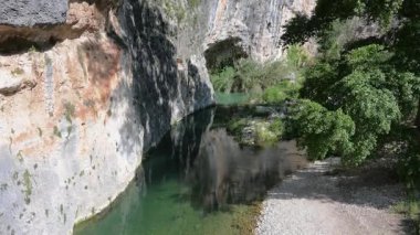 Sakin, berrak nehir, yeşil bitki örtüsü ve çakıllı bir kıyısı olan sarp kayalık bir uçurum boyunca akar ve güneş ışığında sakin bir doğal manzara yaratır..