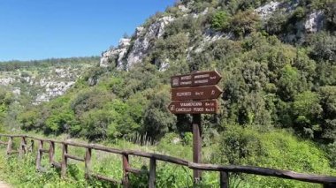 Tahta levhaları ve ahşap çitleri olan tozlu bir yürüyüş yolu uçurumlar, yoğun bitki örtüsü ve parlak mavi bir yay gökyüzü ile çevrili kayalık bir vadi boyunca uzanır..
