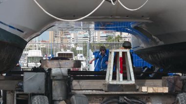 Cagliari - Sardinien - Italien - 03-14-2025: İşçiler, limandaki bir karavan yapısı tarafından desteklenen ve arka planda tekne ve binaların görülebildiği askıya alınmış bir katamaran altında çalışıyorlar.