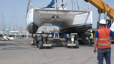 Cagliari - Sardinien - Italien - 03-14-2025: İşçiler limandaki büyük bir vinç kullanılarak yapılan hassas indirme operasyonunda bir katamaranı dikkatlice taşıyıcıya yönlendiriyorlar.