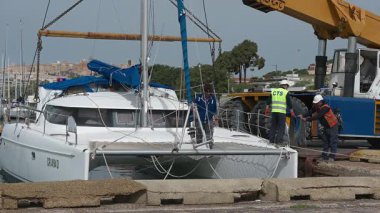 Cagliari - Sardinien - Italien - 03-14-2025: İşçiler, gövdenin altından bir vinç sapanı geçirerek ve kayışları güvenli kaldırma noktalarına koordine ederek bir katamaranı kaldırmaya hazırlanıyorlar.