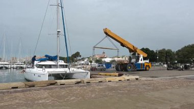 Cagliari - Sardinien - Italien - 03-14-2025: İşçiler ve ekipmanlar limandaki kaldırma operasyonuna hazırlanırken sarı bir vinç beyaz bir katamaranın yakınına doğru hareket ediyor.