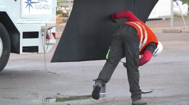 Cagliari - Sardinien - Italien - 03-12-2025: Güvenlik donanımlı bir işçi vinç operasyonundan önce son boyayı bir yelkenlinin omurgasına uygular, destek iskelesinin yakınındaki detayları kontrol eder.