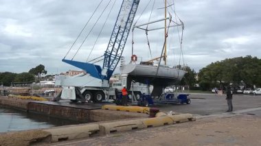Cagliari - Sardinien - Italien - 03-12-2025: Cagliari limanı yakınlarında seyyar bir vinç tarafından bir yelkenli kaldırılırken, mürettebat üyeleri yardım ve bulutlu hava koşullarında gökyüzünü kaplıyor.