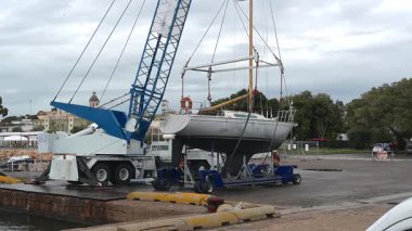 Cagliari - Sardinien - Italien - 03-12-2025: İşçiler bir yelkenliyi hareketli vinç kullanarak tekerlekli bir beşikte konumlandırırken zinciri ayarlar ve gemiyi kaldıracak şekilde stabilize ederler.