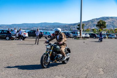 Reggio Calabria - Kalabrien - 05-18-2025: Seçkin beyefendiler Reggio Calabria 'da araba ve tepelerle dolaşırken takım elbiseli motosikletli bir adam otoparkta geziniyor.