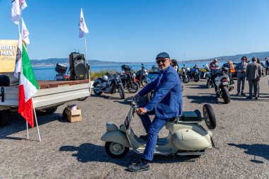 Reggio Calabria - Kalabrien - 05-18-2025: Seçkin Beyefendiler 'de eski mobiletli mavi takım elbiseli adam motosikletlerle, insanlarla ve rıhtımın yanında bayraklarla geziyor.