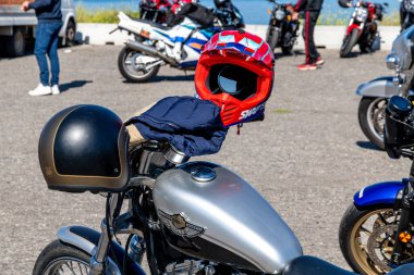 Reggio Calabria - Kalabrien - 05-18-2025: Kırmızı bir motokros kaskı ve siyah bir kask, arka planda park halindeki diğer motosikletlerle birlikte gümüş bir motosikletin üzerinde dinleniyor..