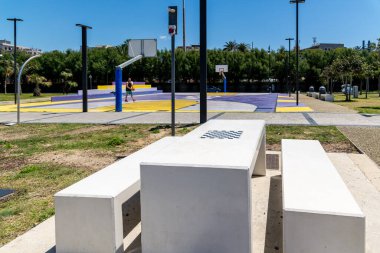 Reggio Calabria - Kalabrien - 05-22-2025: Halka açık bir şehir parkında mor ve sarı bir basketbol sahasının yakınında dahili satranç tahtası bulunan beyaz beton bir piknik masası.