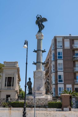 Reggio Calabria - Kalabrien - 05-22-2025: Uzun bir sütunda kanatlı heykeli olan tarihi anıt, parlak mavi bir gökyüzü altında, rakamlar ve binalarla çevrili, kentsel bir ortamda.