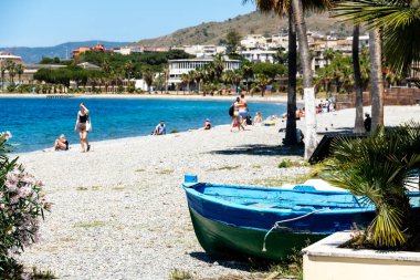 Reggio Calabria - Kalabrien - 05-22-2025: Birkaç kişi gevşeyip turkuaz deniz ve palmiye ağaçlarıyla dolu çakıllı bir plajda yürüyor, ön planda yeşil-mavi bir tekne görünüyor.