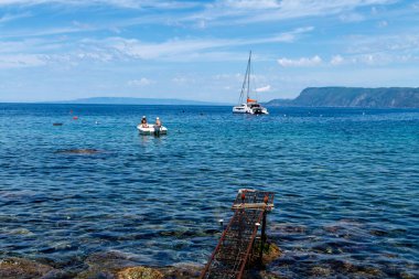 Scilla - Kalabrien - 05-27-2025: İki kişi Scilla, Calabria 'daki dağ kıyısında demirlemiş bir katamaran, ön planda paslı iskeleden geriye kalan.