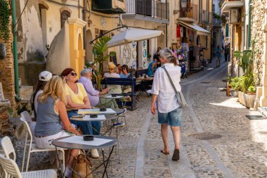 Scilla - Kalabrien - 05-27-2025: İnsanlar Scilla, Calabria 'da bir sokak kafesinde dinlenirken, diğerleri küçük dükkanlar ve konutlarla dolu dar bir sokakta yürüyor.