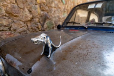 Scilla - Kalabrien - 05-27-2025: Scilla, Calabria 'da, taş bir duvarın yanına park edilmiş eski bir arabanın paslı kaputuna krom bir fare kaputu takılı..