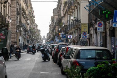 Catania, Sizilien - İtalya - 04-15-2025: Catania 'da park halindeki arabalar, scooterlar, yayalar ve öğleden sonra ışıklarının altındaki mesafeye uzanan tarihi binalarla dolu yoğun bir cadde.