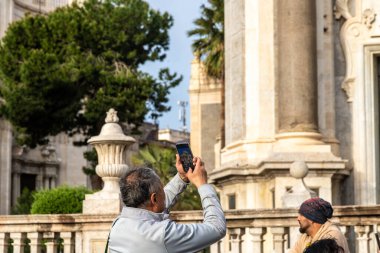 Catania, Sizilien - İtalya - 04-15-2025: Bir adam tarihi taş bir binanın yanında akıllı telefonuyla resim çekiyor, Catania 'da mimari sütunları, balkonu ve yeşillikleri olan bir adam.