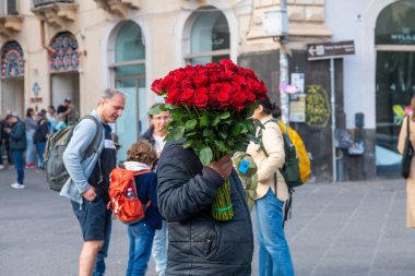 Catania, Sizilien - İtalya - 04-15-2025: Koyu renk ceketli bir çiçekçi Catania, Sicilya 'da kalabalık bir caddede büyük bir buket kırmızı gül tutuyor..