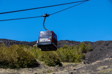 Catania, Sizilien - İtalya - 04-22-2025: Etna tramvayının kırmızı gondolu, Etna Dağı 'nın yamaçlarında açık mavi bir gökyüzü altında yeşil bitki örtüsü ve volkanik arazi üzerinde seyahat ediyor.