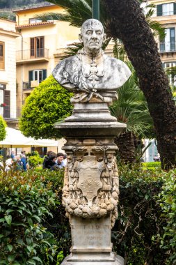 Monreale - Sizilien - Italien - 03-18-2025: Monreale 'de binaları olan bir bahçe meydanında süslü kaide ve yazıtlara sahip dini bir figürün tam mermer heykeli.