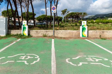 Tropea - Kalabrien - İtalya - 05-29-2025: Tropea, Calabria 'daki elektrikli otomobil şarj istasyonları, yeşil boyalı bir park alanında işaretli semboller, işaretler ve ağaçlar.