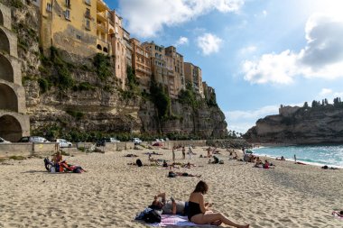 Tropea - Kalabrien - İtalya - 05-29-2025: Sahile gidenler güneşli bir öğleden sonra Tropea 'nın kayalık binalarının altında kumlu sahil boyunca dinlenirler.