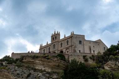 Tropea - Kalabrien - İtalya - 05-29-2025: Tropea, Calabria 'da Santa Maria dellIsola' nın sığınağı, bulutlu gökyüzünün altındaki bir uçurumun üzerinde duruyor ve terasta turistler görülüyor.