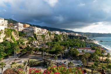 Tropea - Kalabrien - İtalya - 05-29-2025: Calabria 'daki Tropea manzarası kayalık binaları, yemyeşil alanları ve Tyrhenian kıyıları boyunca dramatik bulutların altında bir plajı gösteriyor.