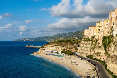 Tropea - Kalabrien - İtalya - 05-29-2025: Tropea, Calabria 'da kumlu bir plajın üzerinde yükselen binalar, burada kumsala gidenler Tyrhenian Denizi boyunca parçalı bulutlu bir gökyüzü altında dinlenirler..