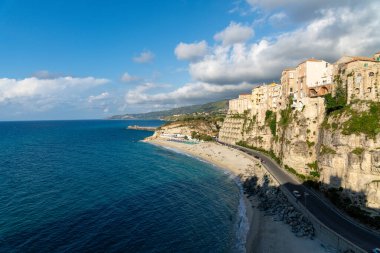 Tropea - Kalabrien - İtalya - 05-29-2025: Calabria 'daki Tropea, kumlu bir sahil üzerindeki tarihi uçurumlar ve parçalı bulutlu bir gökyüzünün altındaki açık mavi deniz üzerindeki tarihi binaları gösteriyor.