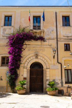 Tropea - Kalabrien - İtalya - 05-30-2025: İtalya 'nın Calabria kentindeki Tropea belediye binasının ön girişi, iki bayrağın altında çiçek açan bougainvillea ve belediye işaretleri barındırıyor.