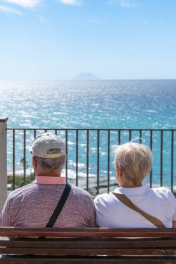 Tropea - Kalabrien - İtalya - 05-30-2025: Tropea, Calabria 'da denize bakan ahşap bir bankta oturan yaşlı bir çift, Stromboli ufuk çizgisinde belli belirsiz bir şekilde görülüyor.