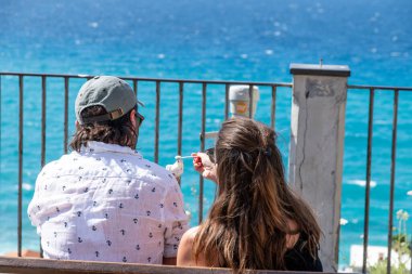 Tropea - Kalabrien - İtalya - 05-30-2025: Bir çift İtalya 'nın Tropea kentinde denize bakan bir bankta otururken, bir kişi diğerine bir kaşık dolusu dondurma sunuyor.