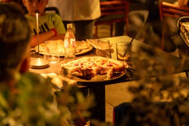 Tropea - Kalabrien - İtalya - 05-30-2025: Birçok kişi akşam bir restoran masasında yemek yiyor, pizza yiyor ve görünür dekor ile sıcak ortam ışıkları altında içiyor.