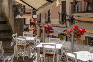 Pizzo - Kalabrien - İtalya - 06-02-2025: Terasında desenli masa örtüleri ve kırmızı çiçekler bulunan küçük açık hava kafesi, balkonları ve tenteleri olan dar bir İtalyan caddesine bakıyor.