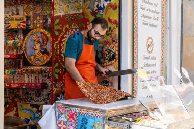Pizzo - Kalabrien - İtalya - 06-02-2025: Turuncu önlüklü tedarikçi, Sicilya 'daki bir şeker dükkanında seramik fayanslar ve şeker vitrinleriyle büyük bir parça renkli fındık kırıntısı kesti.