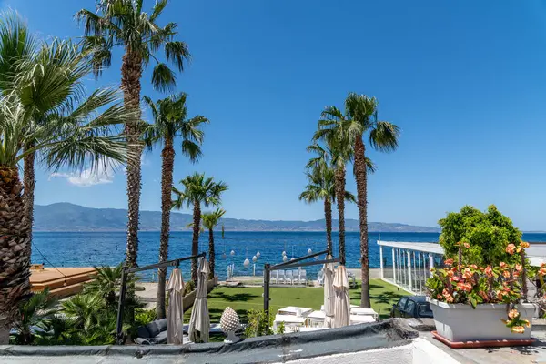 Reggio Calabria - Kalabrien - 05-22-2025: Palmiye ağaçları, güneşlikler ve dinlenme alanları açık bir gökyüzü altında denize ve uzak dağlara bakarak rahatlatıcı bir kıyı atmosferi yaratıyor..