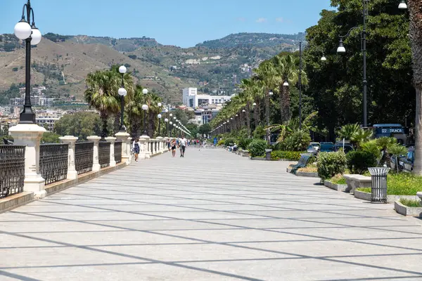 Reggio Calabria - Kalabrien - 05-22-2025: Palmiye ağaçları ve sokak lambalarıyla kaplı geniş bir gezinti alanı, arka planda dağlar, ağaçlar ve şehir binaları yer alıyor.