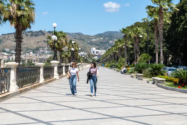 Reggio Calabria - Kalabrien - 05-22-2025: Güneşli bir günde, arka planda banklar, sokak lambaları, ağaçlar ve tepelerle palmiye çizgili bir sahil geçidi boyunca yürüyen iki kadın.