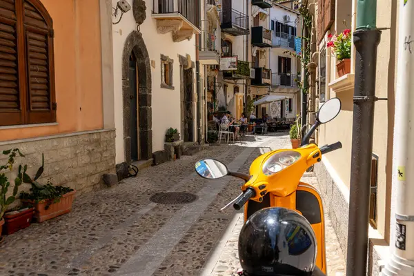 Scilla - Kalabrien - 05-27-2025: Scilla, Calabria 'da dar bir kaldırıma park etmiş kasklı turuncu scooter, arka planda açık bir kafede oturan insanlar.
