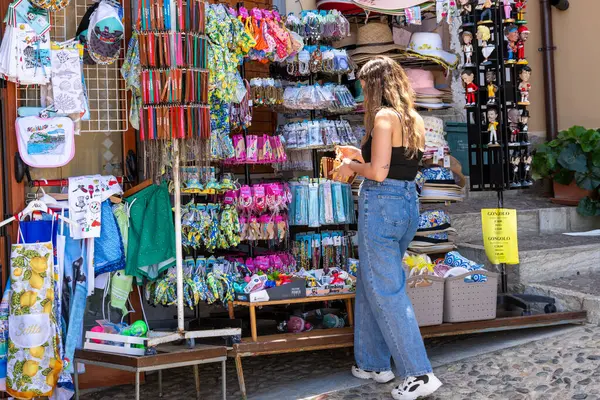 Scilla - Kalabrien - 05-27-2025: Genç bir kadın Scilla, Calabria 'daki renkli bir hediyelik eşya standında mıknatıslar, şapkalar, havlular ve önlükler sergileniyor