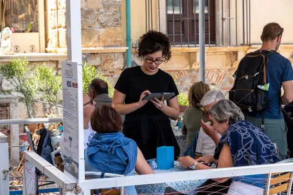 Scilla - Kalabrien - 05-27-2025: Siyahlı bir garson, misafirler gölgeli bir açık kafe masasında içki ve görünür menülerle otururken tabletle sipariş alıyor.