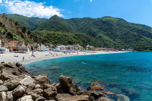 Scilla - Kalabrien - 05-27-2025: Temiz turkuaz suyu, güneşlenen insanlar, beyaz binalar ve Scilla, Kalabrien 'deki yemyeşil tepeler.