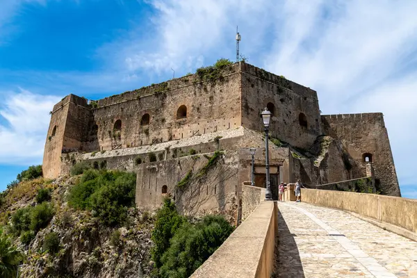 Scilla - Kalabrien - 05-27-2025: Pembe sırt çantalı beyaz elbiseli bir kadın denize bakan bir kalenin yanında selfie çekiyor, arka planda görünen bir ada var..