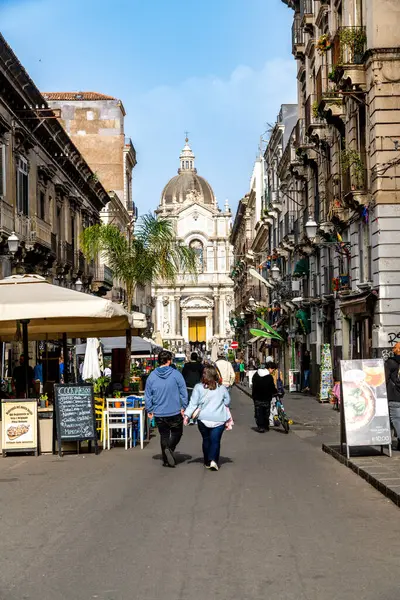 Catania, Sizilien - İtalya - 04-15-2025: Catania, Sicilya 'da restoranları, açık masaları, yayaları ve kubbesi mavi gökyüzünün altında olan barok kilisesi olan işlek cadde.