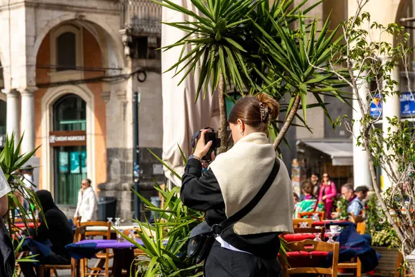 Catania, Sizilien - İtalya - 04-15-2025: Bir kadın Catania 'da bir açık kafe yakınında fotoğraf çekiyor, çerçevelenmiş palmiye bitkileri ve insanlarla çevrili ve tarihi mimariyle çevrili.