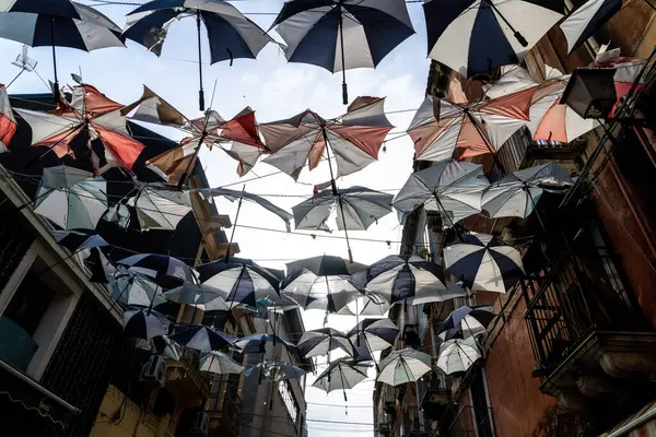 Catania, Sizilien - İtalya - 04-15-2025: Düzinelerce renkli şemsiye Catania 'da dar bir caddenin üzerinde asılı duruyor, yüksek binalar arasında, çarpıcı bir tepe örtüsü kurulumu yaratıyor.