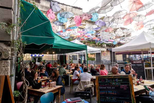 Catania, Sizilien - İtalya - 04-15-2025: Catania 'daki açık hava restoranı sahnesi, masaların üstündeki kablolara asılmış renkli şemsiyeler ve görünür yemek menüleri ile insanların yemek yediği yer..