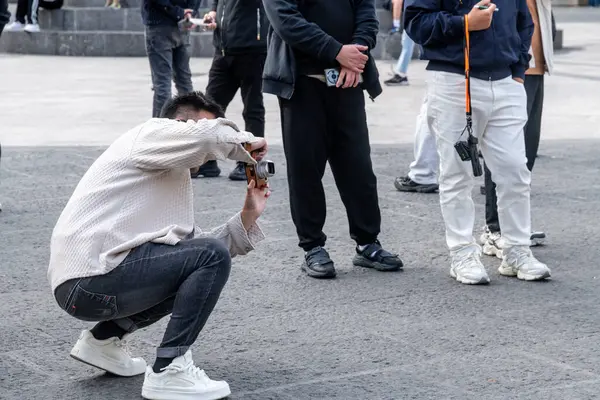 Catania, Sizilien - İtalya - 04-15-2025: Bir halk meydanında sıradan giyinmiş insanlarla çevrili, fotoğraf çekilirken taş kaldırımda çömelmiş bir adam.