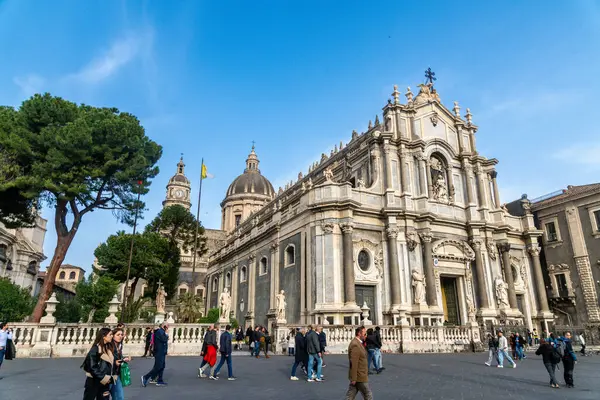 Catania, Sizilien - İtalya - 04-15-2025: Catania 'daki SantAgata Katedrali' nin heykelleri, kubbeleri, saat kulesi, ağaçları ve halk meydanında yürüyen insanların yan perspektifi.