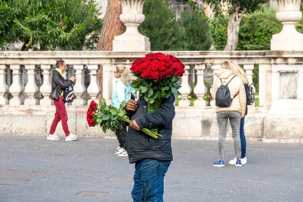 Catania, Sizilien - İtalya - 04-15-2025: Çiçekçi elinde iki büyük buket kırmızı gülle, yüzünü kısmen gizliyor, yayalar Catania 'da bir kayanın yanından geçerken,.